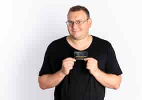 Free photo overweight man in glasses wearing black t-shirt showing credit card looking at camera with happy face standing over white wall