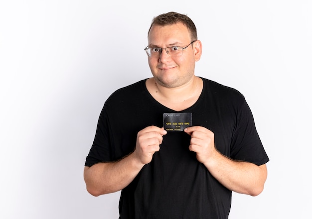 Overweight man in glasses wearing black t-shirt showing credit card looking at camera with happy face standing over white wall