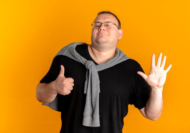 Overweight man in glasses wearing black t-shirt showing and pointing up with fingers number six standing over orange wall