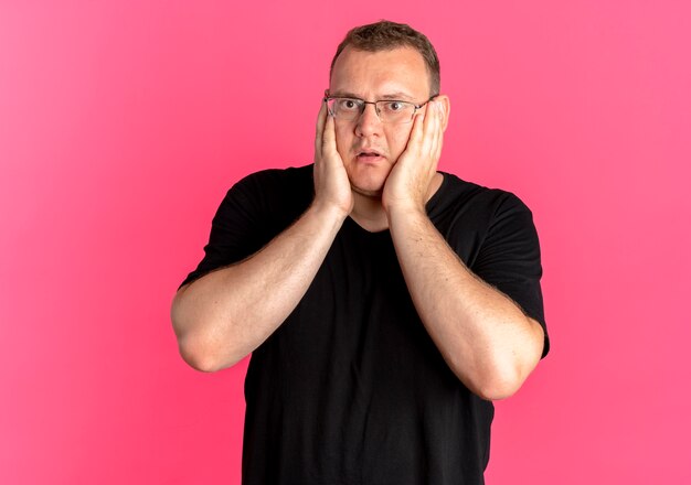 Overweight man in glasses wearing black t-shirt surprised and amazed over pink