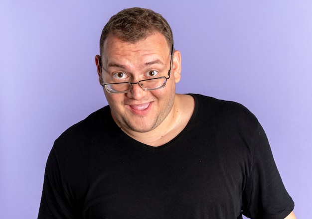 Free photo overweight man in glasses wearing black t-shirt surprised and happy over blue