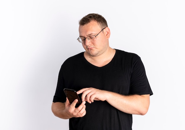Полный мужчина в очках, одетый в черную футболку, переписывается с кем-то с помощью смартфона, стоящего у белой стены