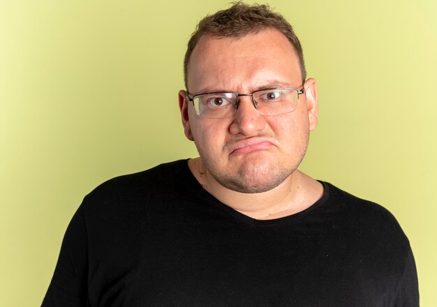 Overweight man in glasses wearing black t-shirt with frowning face displeased over light