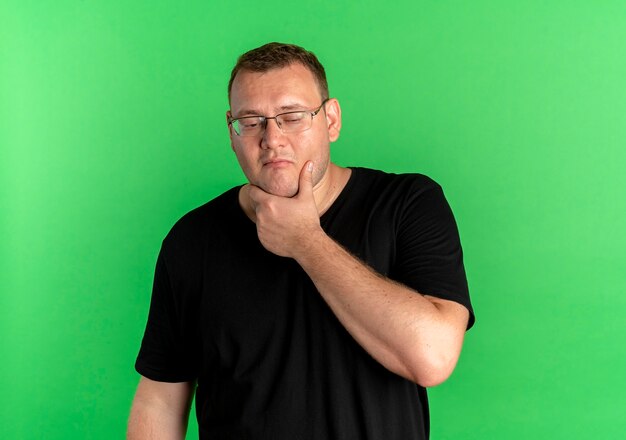 녹색 벽 위에 서있는 얼굴에 잠겨있는 표정으로 턱에 손으로 검은 티셔츠를 입고 안경에 과체중 남자