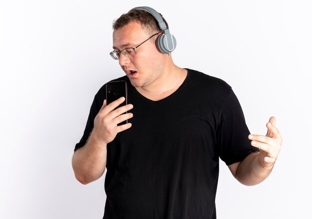 흰 벽 위에 서있는 마이크 노래로 자신의 스마트 폰을 사용하는 헤드폰으로 검은 색 티셔츠를 입고 안경에 과체중 남자