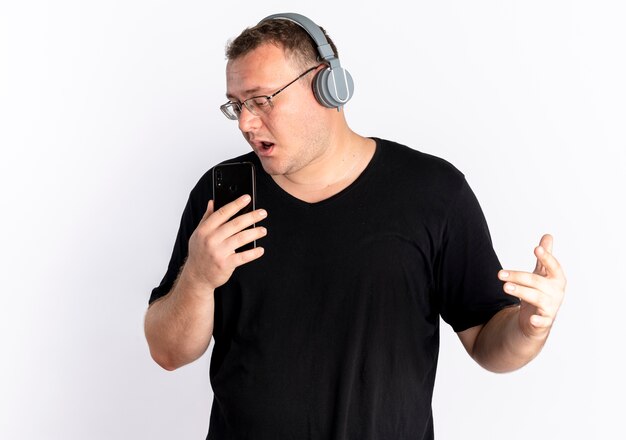 Overweight man in glasses wearing black t-shirt with headphones using his smartphone as microphone singing standing over white wall