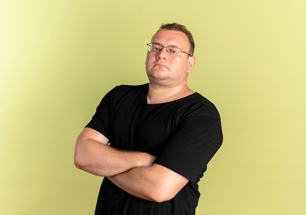 Free photo overweight man in glasses wearing black t-shirt  with seriosu confident expression with arms crossed on chest standing over light wall