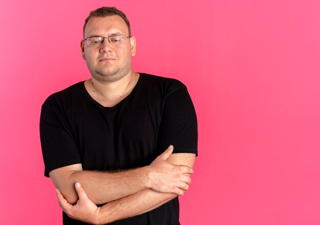 Overweight man in glasses wearing black t-shirt  with smile on face with arms crossed on chest standing over pink wall