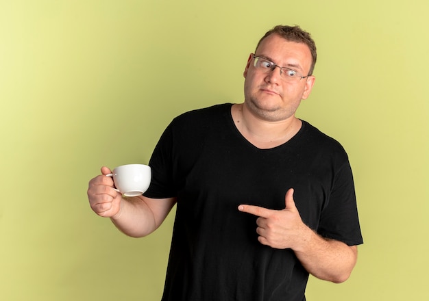 Бесплатное фото Полный мужчина в очках, одетый в черную футболку, держит чашку кофе, указывая пальцем на нее, смущенно глядя в сторону, стоя над светлой стеной