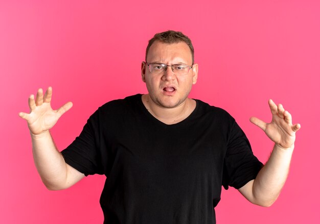 無料写真 ピンクの壁の上に立って混乱し、不快に見える黒いtシャツを着た眼鏡の太りすぎの男