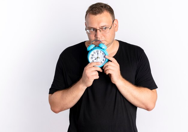 Overweight man wearing black t-shirt in glasses holding alarm clock looking aside with sad expression standing over white wall