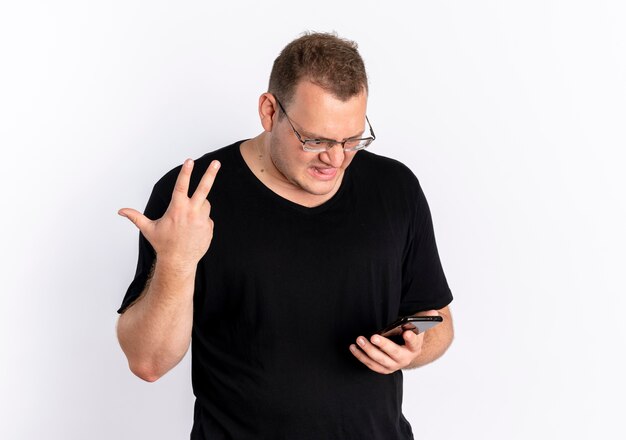 眼鏡をかけた黒のTシャツを着てスマートフォンの画面を見ている太りすぎの男性は、白い壁の上に立っている手でジェスチャーを混乱させ、不快に感じました