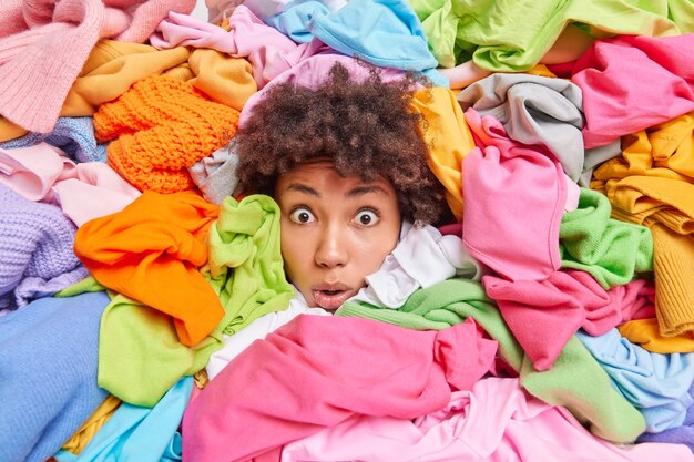 Overwhelmed Afro American woman gives advice to recycle your old clothes sticks out head through multicolored clothing surrouunded by unwearable items collected for donation. Textile recycling