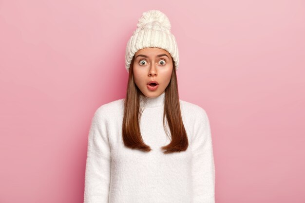 Overwhelmed astonished woman keeps mouth opened from fear, gasps amazed, stares at camera, wears white hat with pompon and sweater, isolated over pink background