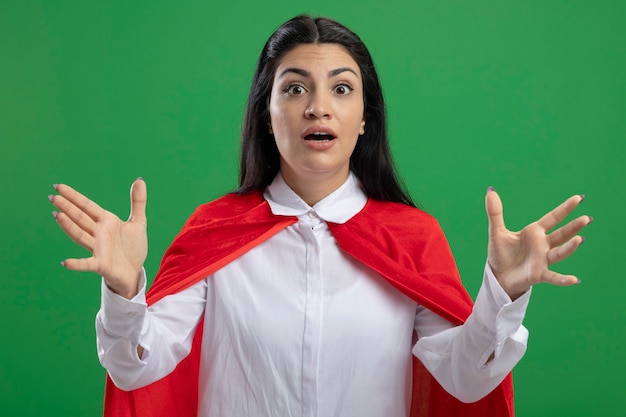 Overwhelmed young caucasian superhero girl spreading hands with her mouth open isolated on green wall