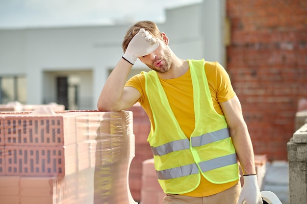 Overwork. Tired man in bright vest taking off his protective helmet and touches face with hand standing near pile of bricks at construction site on fine day