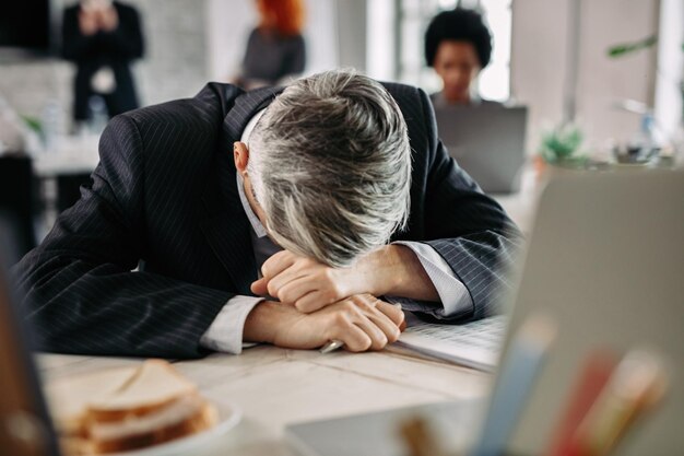 과로한 사업가는 사무실에서 피곤함을 느끼고 책상에서 머리를 쉬고 있다 배경에 사람들이 있다