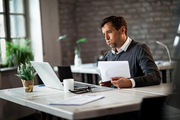 Overworked businessman using laptop while going through business reports and working in the office