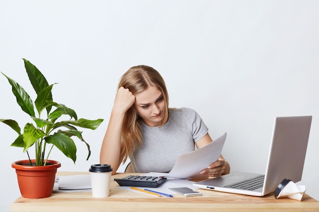 Overworked businesswoman sitting at wooden table, surrounded with modern gadgets, reading documents attentively, trying to understand everything. Female accounter calculating bills and expenses