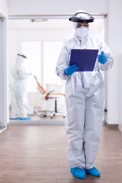 Overworked doctor with suits against contamination with coronavirus writing notes on clipboard. Medical personal dressed in protection equipment against infection with covid-19 during global pandemic.