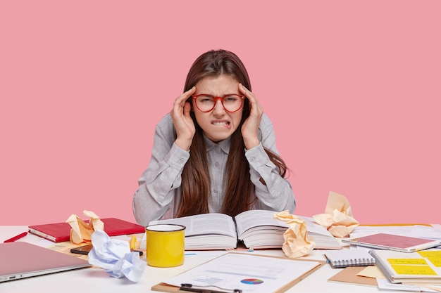 Overworked pretty woman bites lower lip, suffers from headache, reads scientific article in book, has mess on table