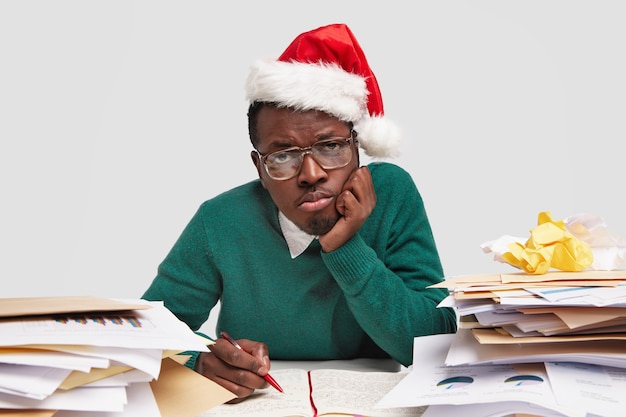 働き過ぎの悲しいアフリカ系アメリカ人の男は頬に手を保ち、サンタクロースの帽子、光学眼鏡をかけ、日記に書き留めます