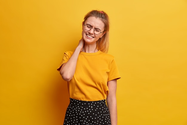 Free photo overworked schoolgirl feels pain in back after sitting long time at computer during distance learning, touches neck, tilts head, frowns from pain