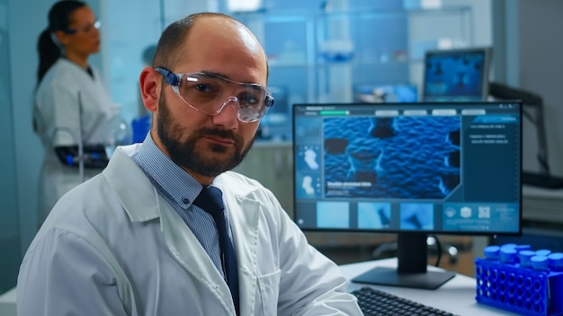 Overworked scientist with protection glasses looking at camera sighing sitting in research laboratory. Doctors examining virus evolution using high tech and chemistry tools for medical research.