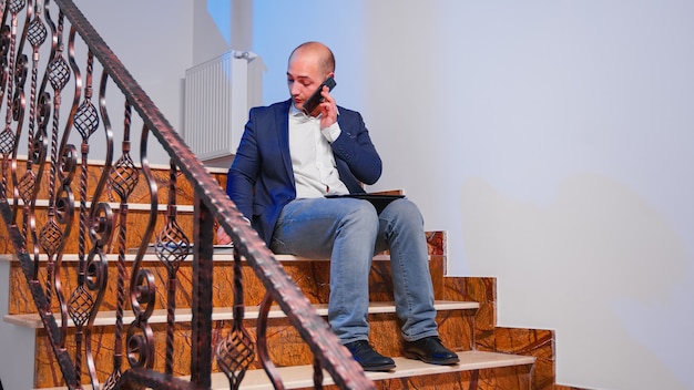 Overworked tired businessman reading project deadline during phone call with corporate manager. Serious entrepreneur working on job sitting on staircase of business building late at night.