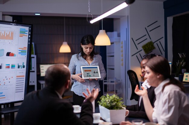 Overworked workaholic entrepreneur woman shwoing marketing graphs using tablet overworking at company solution late at night in meeting room