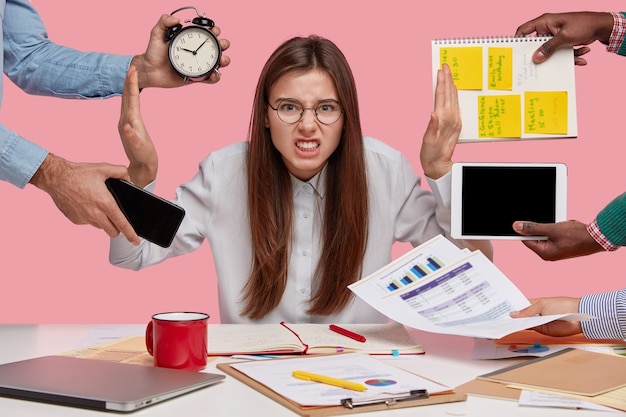 Free photo overworked young employee refuses all things, frowns face in annoyance, sits at desktop with paper documents and notepad, isolated over pink wall. female worker bothered by many questions