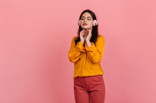 Pacified dark-haired lady in yellow shirt and bright trousers enjoys classical music in headphones on pink wall.