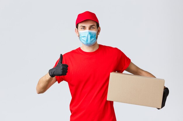 Packages and parcels delivery, covid-19 quarantine delivery, transfer orders. Cheerful courier in red uniform, gloves and face mask, thumb-up, recommend contactless deliver, holding box with order