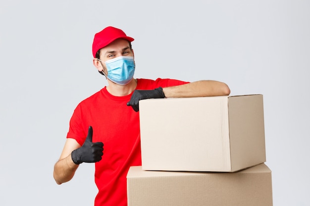 Packages and parcels delivery, covid-19 quarantine and transfer orders. Confident courier in red uniform, gloves and medical mask, encourage call service, show thumb-up lean on boxes