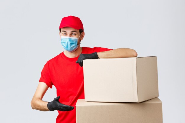 Packages and parcels delivery, covid-19 quarantine and transfer orders. Smiling courier in red uniform, gloves and medical face mask, introduce boxes to transfer your order, recommend service