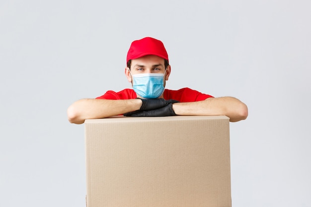 Packages and parcels delivery, covid-19 quarantine and transfer orders. Young courier in red uniform cap, face mask and gloves, leaning on box to deliver, shipping your orders, grey background