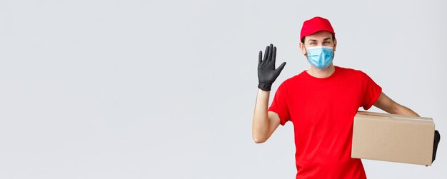 Packages and parcels delivery covid19 quarantine delivery transfer orders Friendly courier in red uniform face mask with protective gloves deliver order box to client waving hand in hello