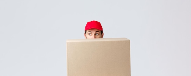 Packages and parcels delivery covid19 quarantine and transfer orders Scared courier in red uniform cap hiding behind customer order looking left nervously peeking at banner or advertisement