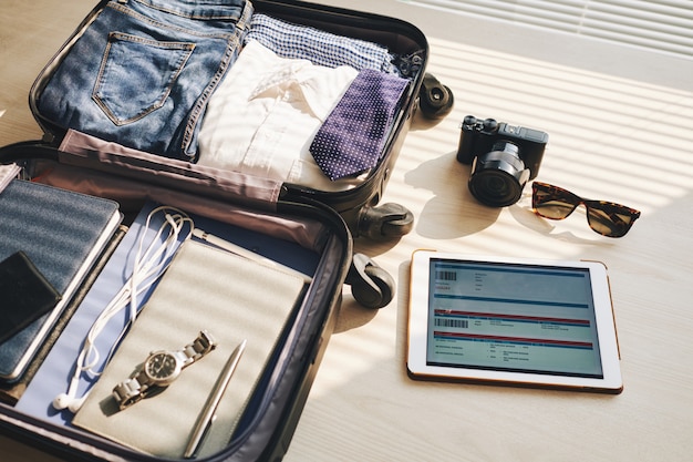 Packed suitcase on desk, tablet with eticket on screen, camera, and sunglasses