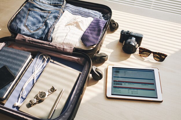 Packed suitcase on desk, tablet with eticket on screen, camera, and sunglasses