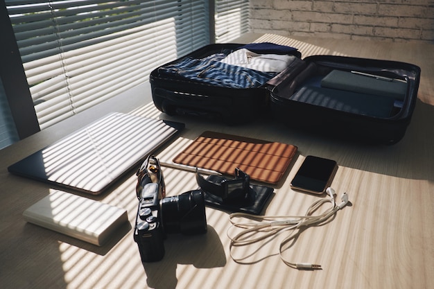 Упакованный чемодан, лежащий на столе у окна с жалюзи, и электронные устройства поблизости