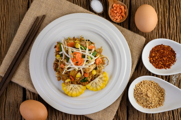 Pad thai in a white plate with lemon, eggs and seasoning on a wooden table.