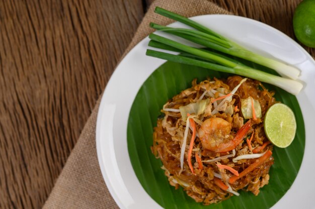 Padthai shrimp in a white dish with lime and eggs on wooden table.