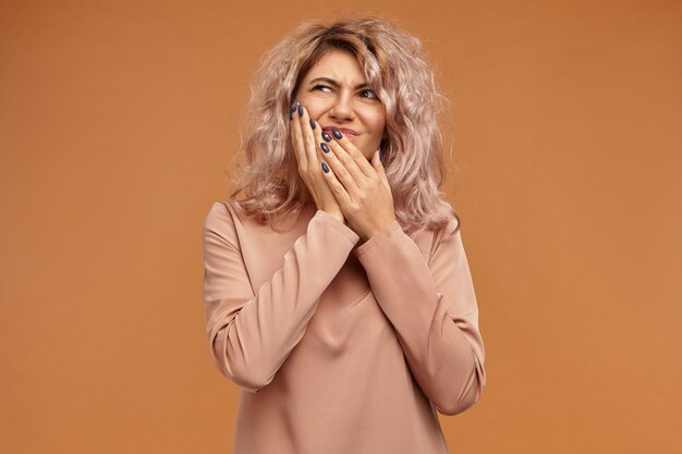 Pain, illness, health, sickness, dental care, people and lifestyle concept. Emotional frustrated young Caucasian woman with voluminous hairdo putting hand on her cheek