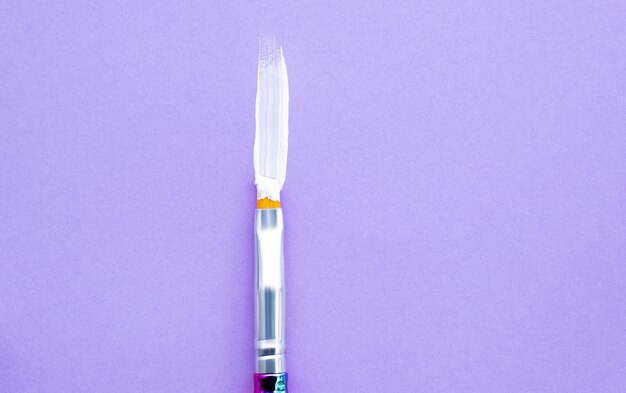 Paint brush with white acrylic paint on purple background.