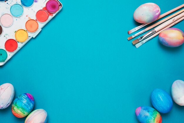 Paint brushes; easter eggs and colorful watercolor box on blue backdrop with space in the center