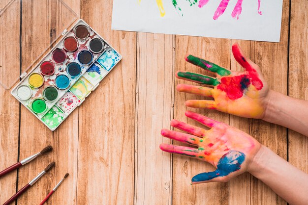 Painted hands with watercolor palette; brushes and paper on wooden table