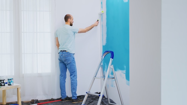 Painting blue wall with white paint using roller brush during home renovation. Handyman renovating. Apartment redecoration and home construction while renovating and improving. Repair and decorating.