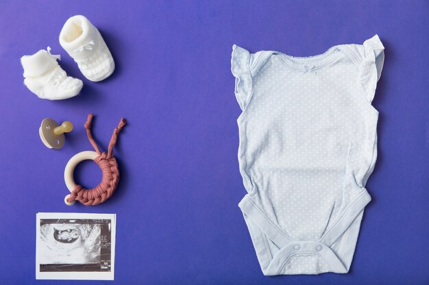 Pair of baby woolen shoes; pacifier; toy; baby onesie and ultrasound picture on blue background