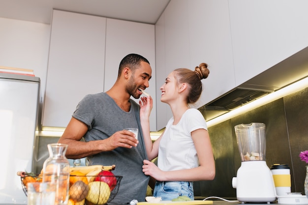 Pair flirting in the kitchen and showing their love. Wife gives her husband to try a piece of fruit, keeps his T-shirt. Couple with passion and happiness looking at each other. Fans of a healthy diet.